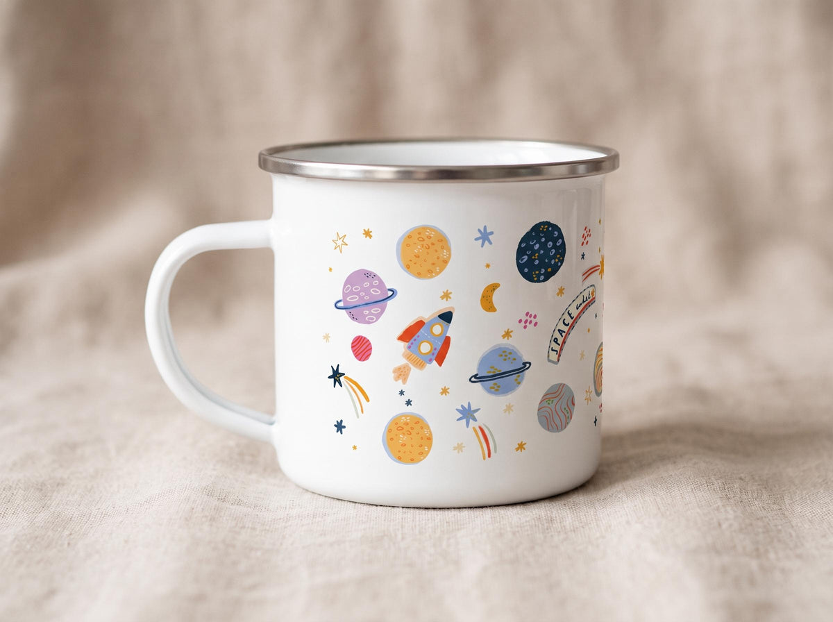 A white enamel mug with a silver rim and a handle, featuring a colourful space-themed pattern of planets, stars, a rocket ship, and a banner with the text SPACE AWAITS in a playful font, set against a neutral-toned fabric background.