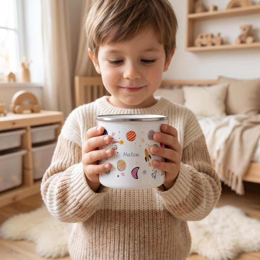 A white enamel mug with a silver rim featuring a colourful space-themed pattern of planets, stars, and a rocket ship, with the name Mateo printed in the centre. The mug is held by two hands, showcasing the detailed celestial graphic design.