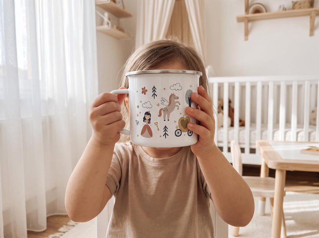 A white enamel mug with a silver rim featuring a printed pattern of a princess, a winged horse, a pumpkin carriage, trees, clouds, and a key. The mug has a side handle and is held up to obscure the face of the person holding it.