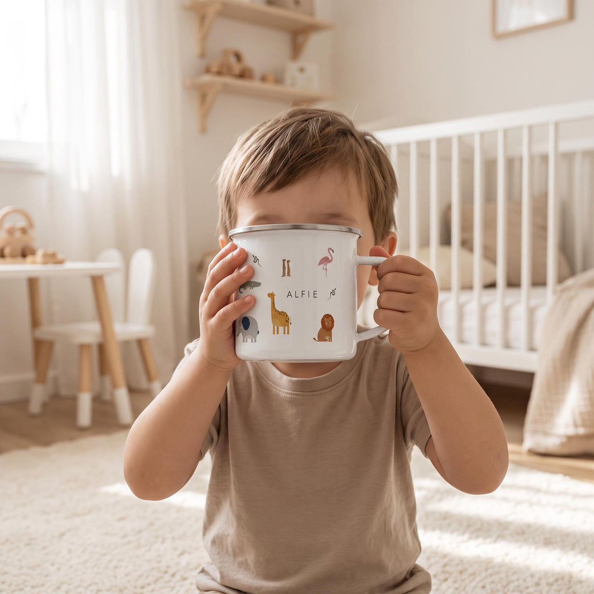 A white enamel mug with a silver rim and handle, featuring colourful illustrations of various animals including a giraffe, elephant, lion, and flamingo, with the name ALFIE printed in black capital letters in the centre.