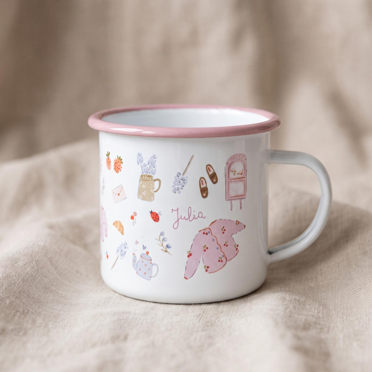 A white enamel mug with a pink rim and handle, featuring a variety of small, colourful illustrations including a pink cardigan, a teapot, a mailbox, flowers, and the name Julia written in cursive script across the centre of the mug.