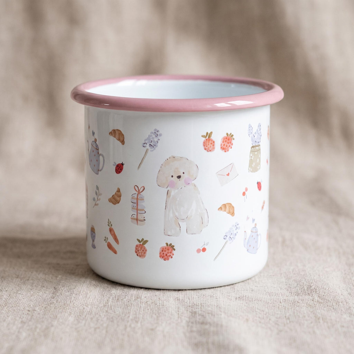 A white enamel mug with a pink rim featuring a repeating pattern of watercolour illustrations including a small dog, croissants, strawberries, carrots, teapots, ladybugs, envelopes, and floral sprigs scattered across the exterior surface.