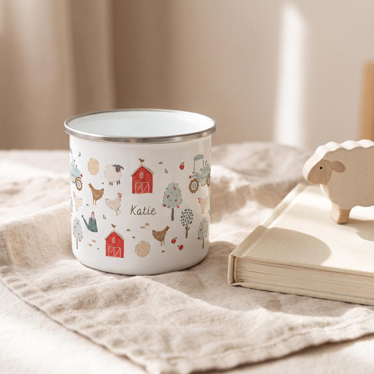 A white enamel mug with a silver rim featuring a colourful farm-themed pattern of red barns, tractors, chickens, sheep, trees, and apples. The name Katie is printed in the centre of the design among the various farm illustrations.