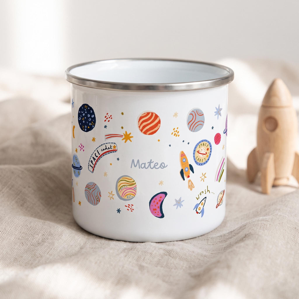 A white enamel mug with a silver rim featuring a colourful space-themed pattern of planets, stars, rockets, and the text "Space cadet" and "Mateo" printed on the side. The design includes various celestial shapes in orange, blue, and pink.