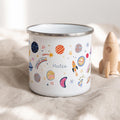 A white enamel mug with a silver rim featuring a colourful space-themed pattern of planets, stars, rockets, and the text "Space cadet" and "Mateo" printed on the side. The design includes various celestial shapes in orange, blue, and pink.