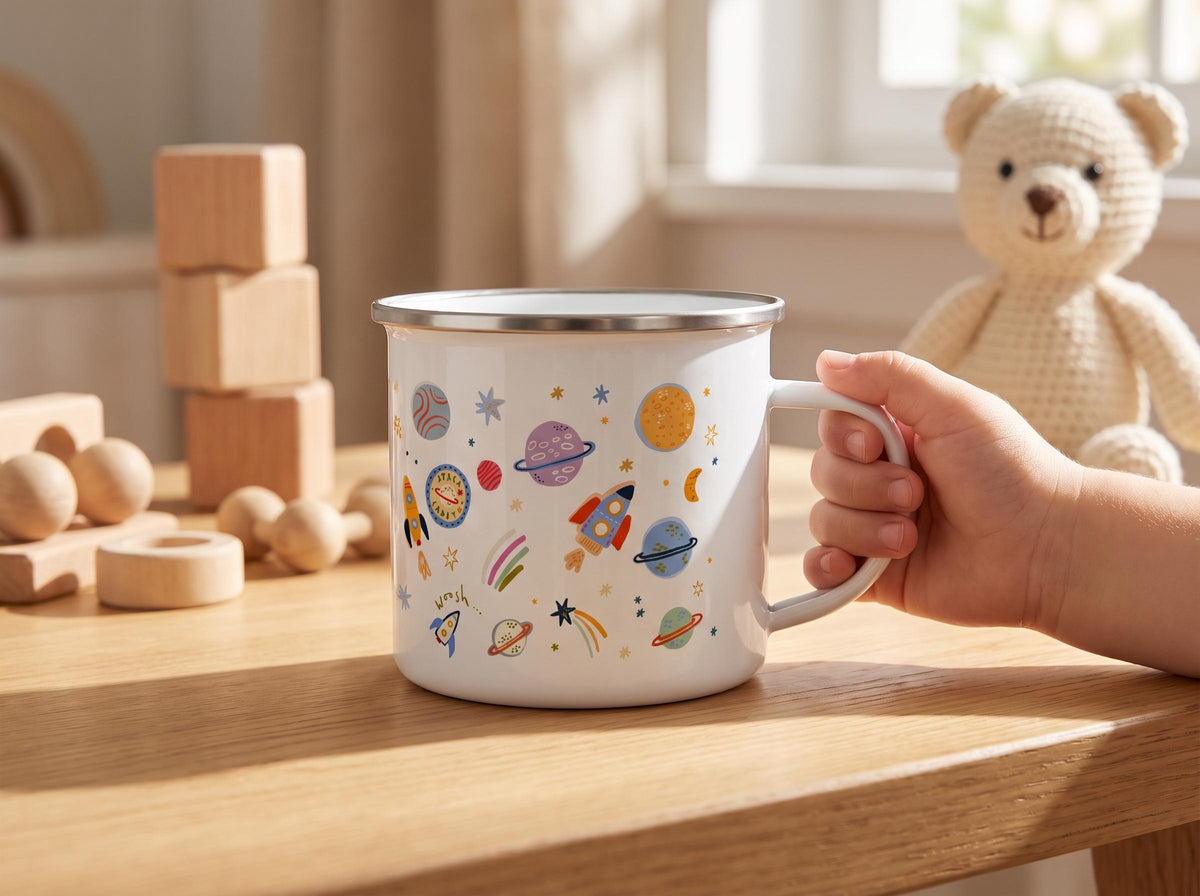 A white enamel mug featuring a colourful pattern of rockets, planets, stars, and moons in shades of orange, blue, and purple. The mug has a silver-toned rim and a sturdy handle, displaying a playful space-themed design on its exterior.