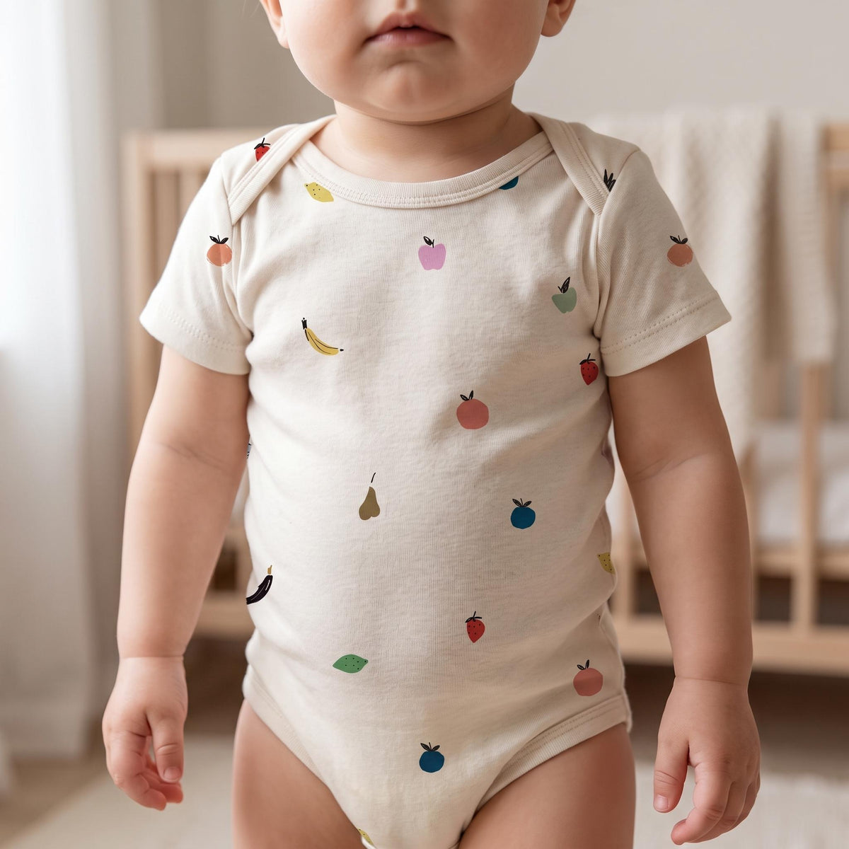 A short-sleeved baby bodysuit in a light cream colour featuring a scattered pattern of small, colourful fruit illustrations including bananas, strawberries, apples, pears, and blueberries distributed across the front of the garment.