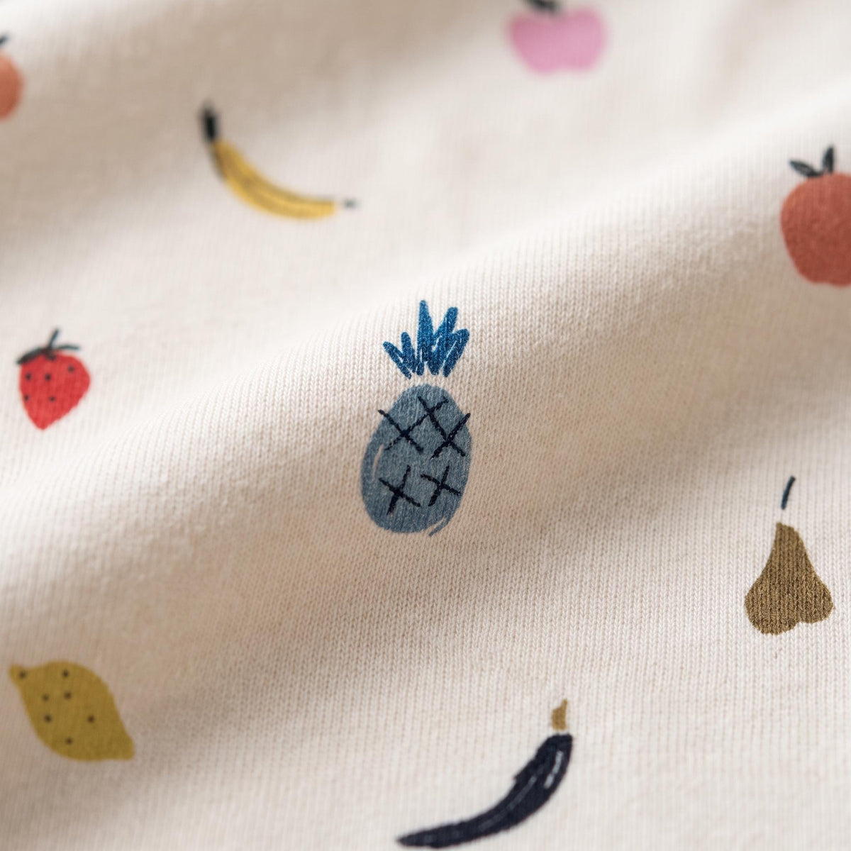 A cream-coloured fabric featuring a repeating pattern of small, hand-drawn style fruit illustrations including a blue pineapple, a red strawberry, a yellow banana, a brown pear, a pink apple, and a yellow lemon scattered across the surface.