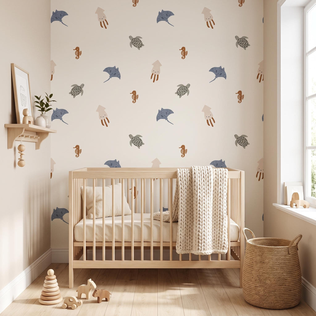 A light-coloured wooden cot with a cream-coloured chunky knit blanket and a matching pillow. The wall behind features a repeating pattern of sea creatures including blue stingrays, brown seahorses, and grey turtles on a beige background.