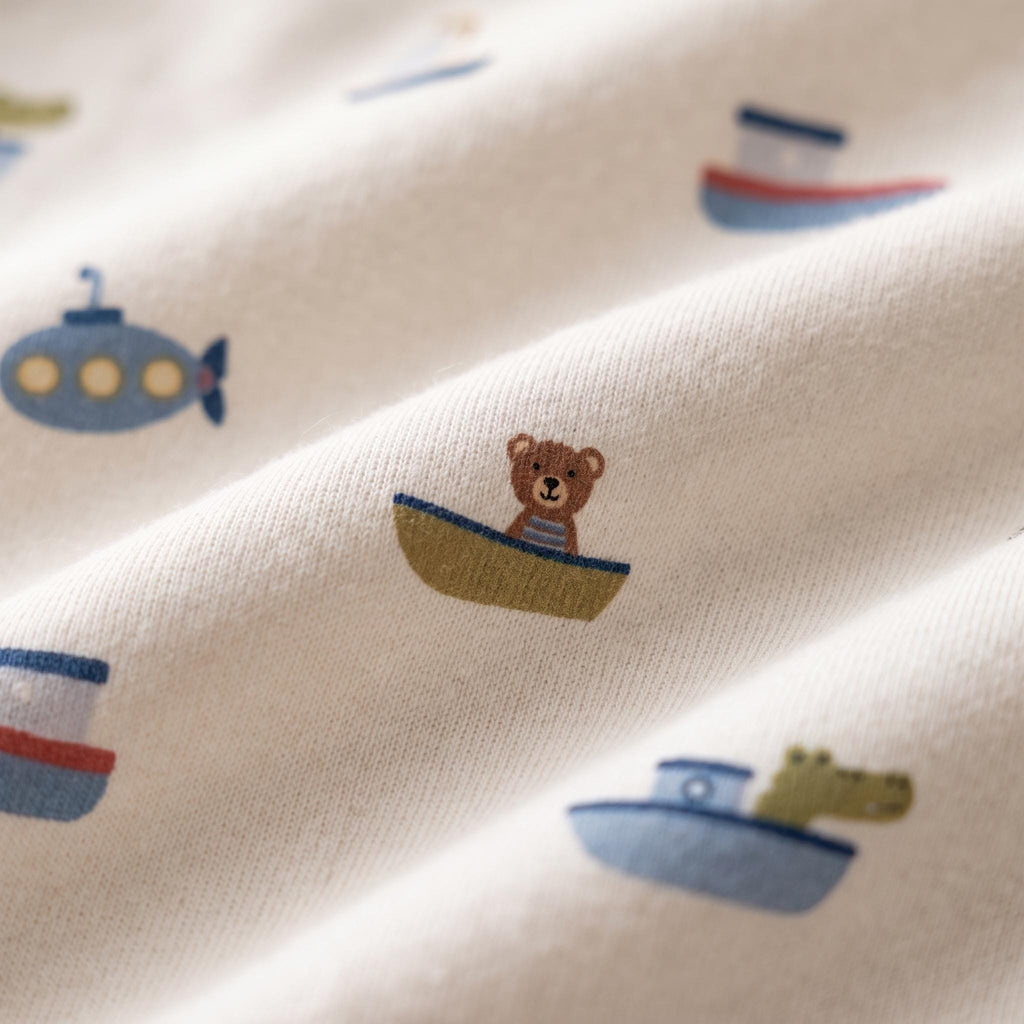 A cream-coloured fabric featuring a repeating pattern of small illustrations including a brown bear in a green boat, blue submarines, red and blue boats, and a green crocodile in a blue boat, all printed in a soft, matte finish.