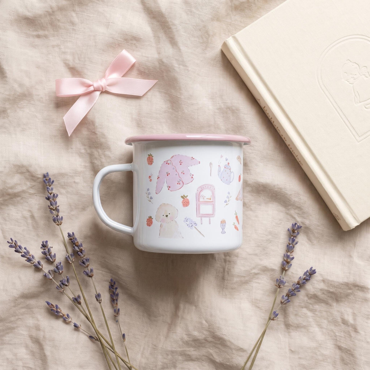 A white enamel mug with a pink rim featuring a repeating pattern of small illustrations including a white dog, a pink cardigan, a teapot, carrots, and strawberries. The mug is accompanied by a light pink ribbon and dried lavender.