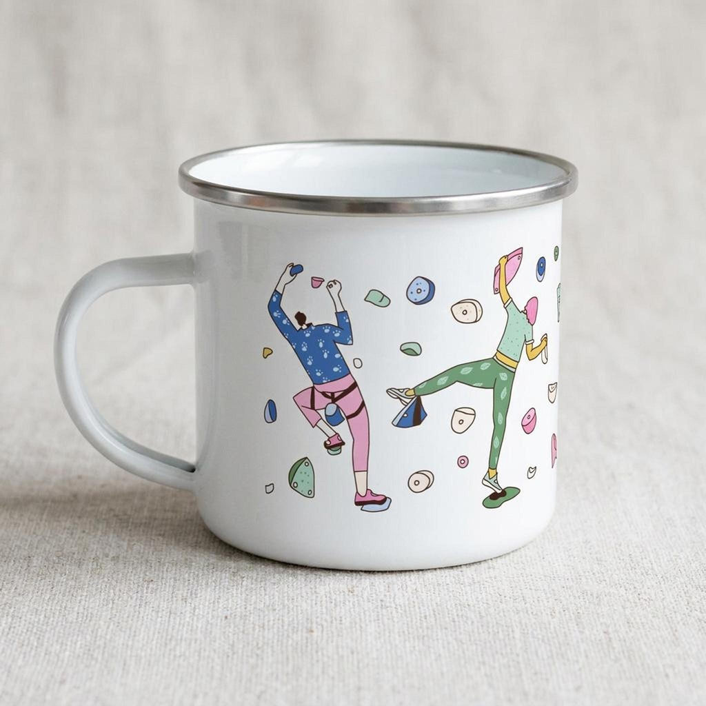 A white enamel mug with a colourful rock climbing pattern stands on fabric.