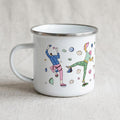A white enamel mug with a colourful rock climbing pattern stands on fabric.