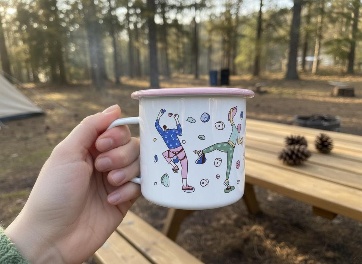 A hand holds a white enamel mug with pink trim in a wooded campsite setting.
