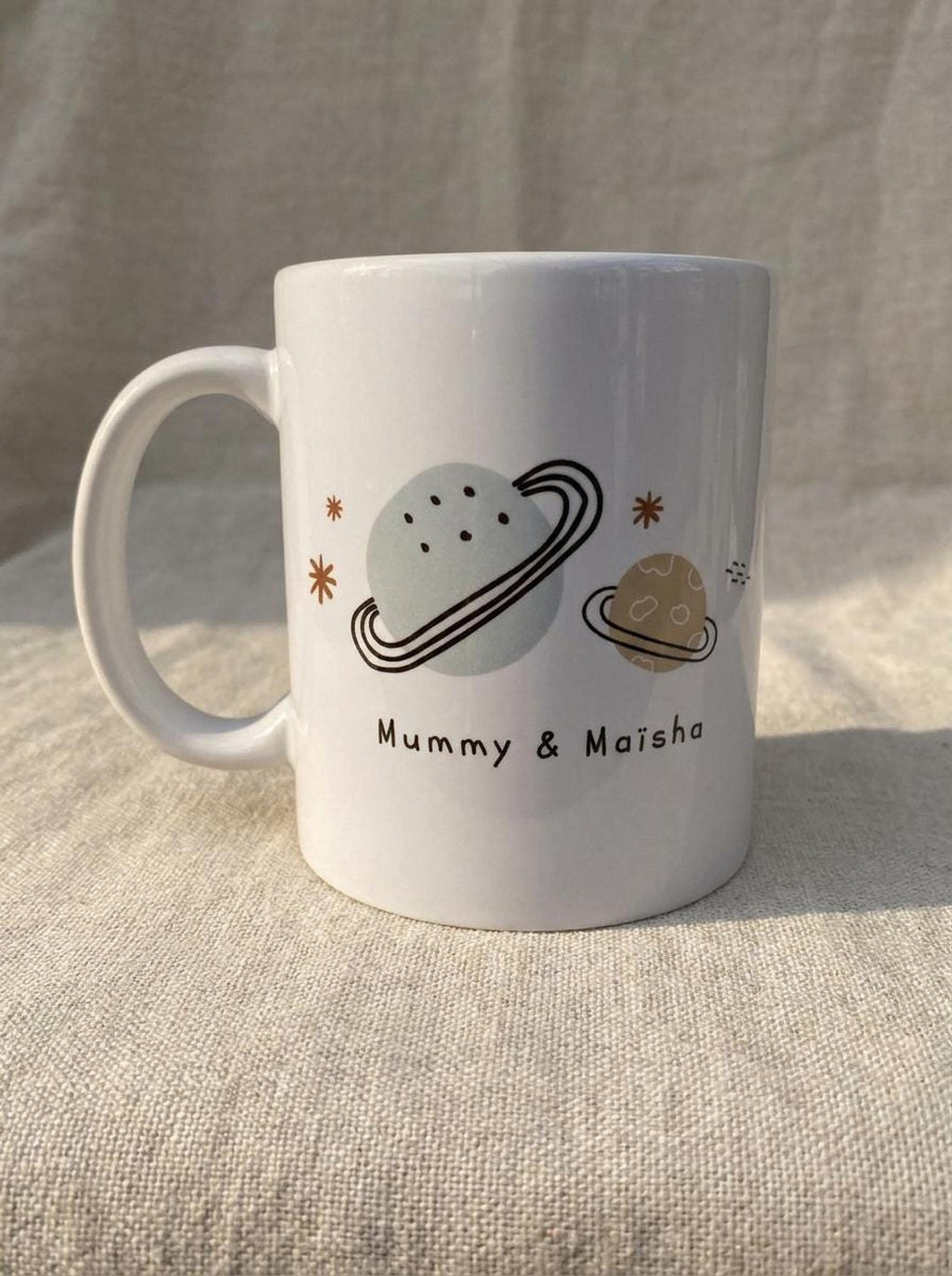 A single large white ceramic mug is printed with illustrations of planets, stars, and the text Mummy & Maisha.