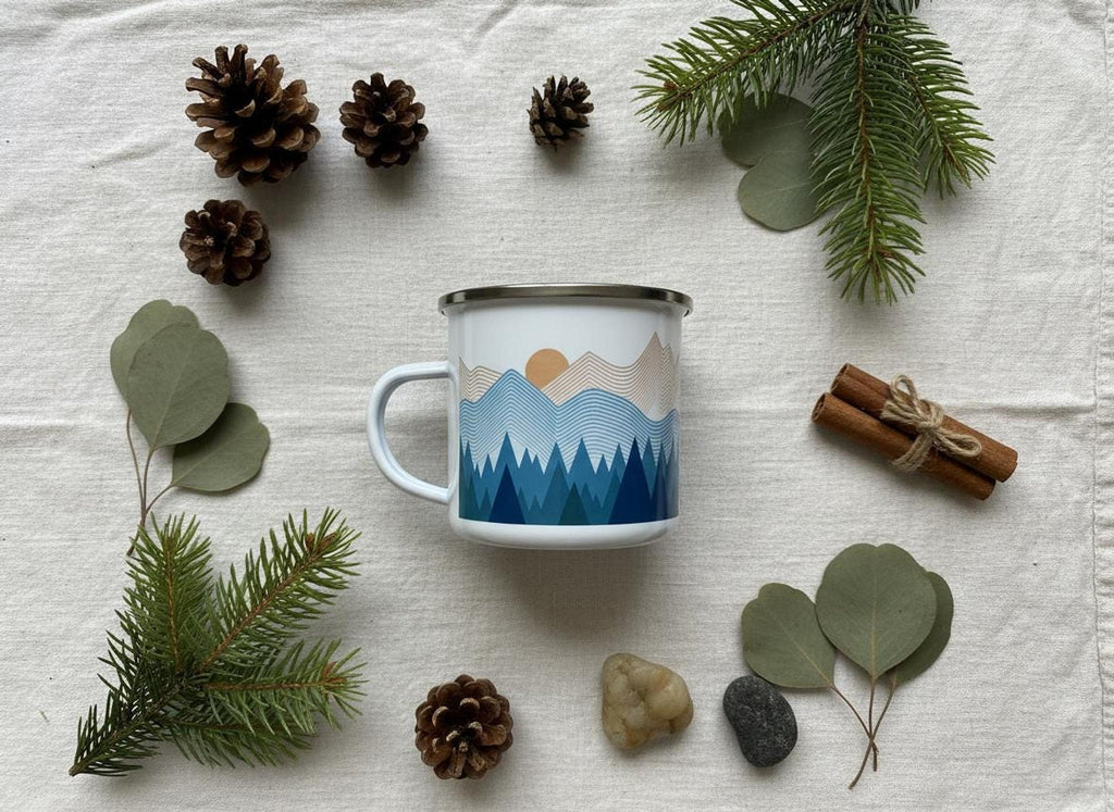A white enamel mug featuring a blue mountain design lies flat on a light fabric surface surrounded by pine cones, evergreen sprigs, and eucalyptus leaves.