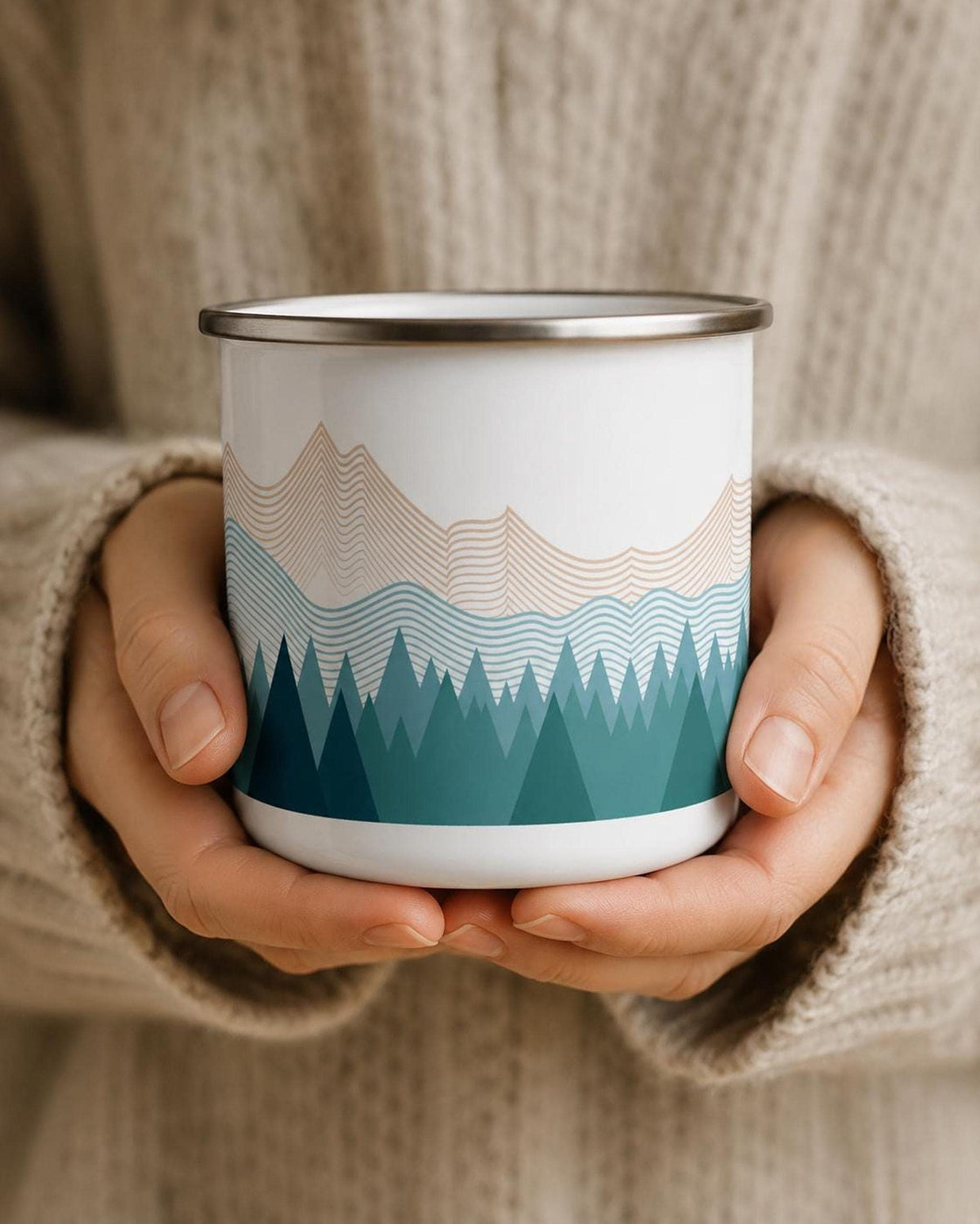 A person wearing a cream sweater holds a white enamel mug displaying a teal pine tree and striped mountain range design.