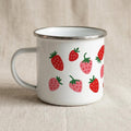 A white metal enamel mug with a silver rim and handle sits on a light fabric background, featuring a pattern of red and pink strawberries.