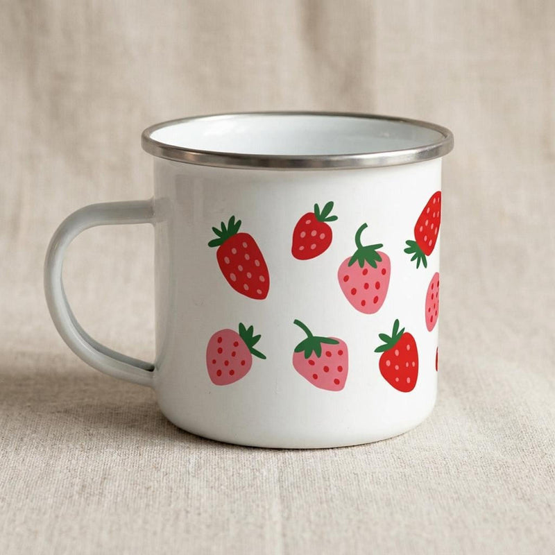 A white metal enamel mug with a silver rim and handle sits on a light fabric background, featuring a pattern of red and pink strawberries.