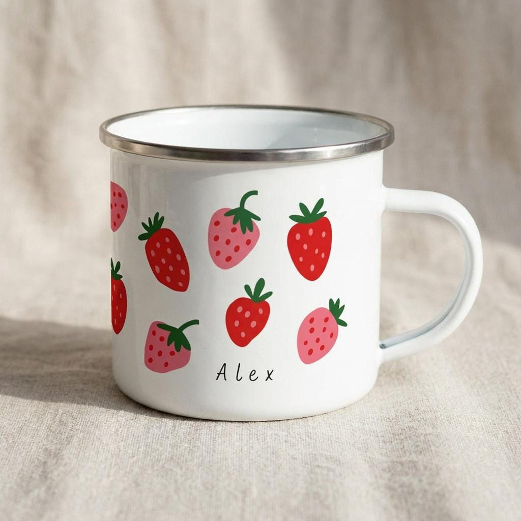 A white enamel mug featuring a strawberry pattern and the custom name Alex is displayed on a textured fabric surface.