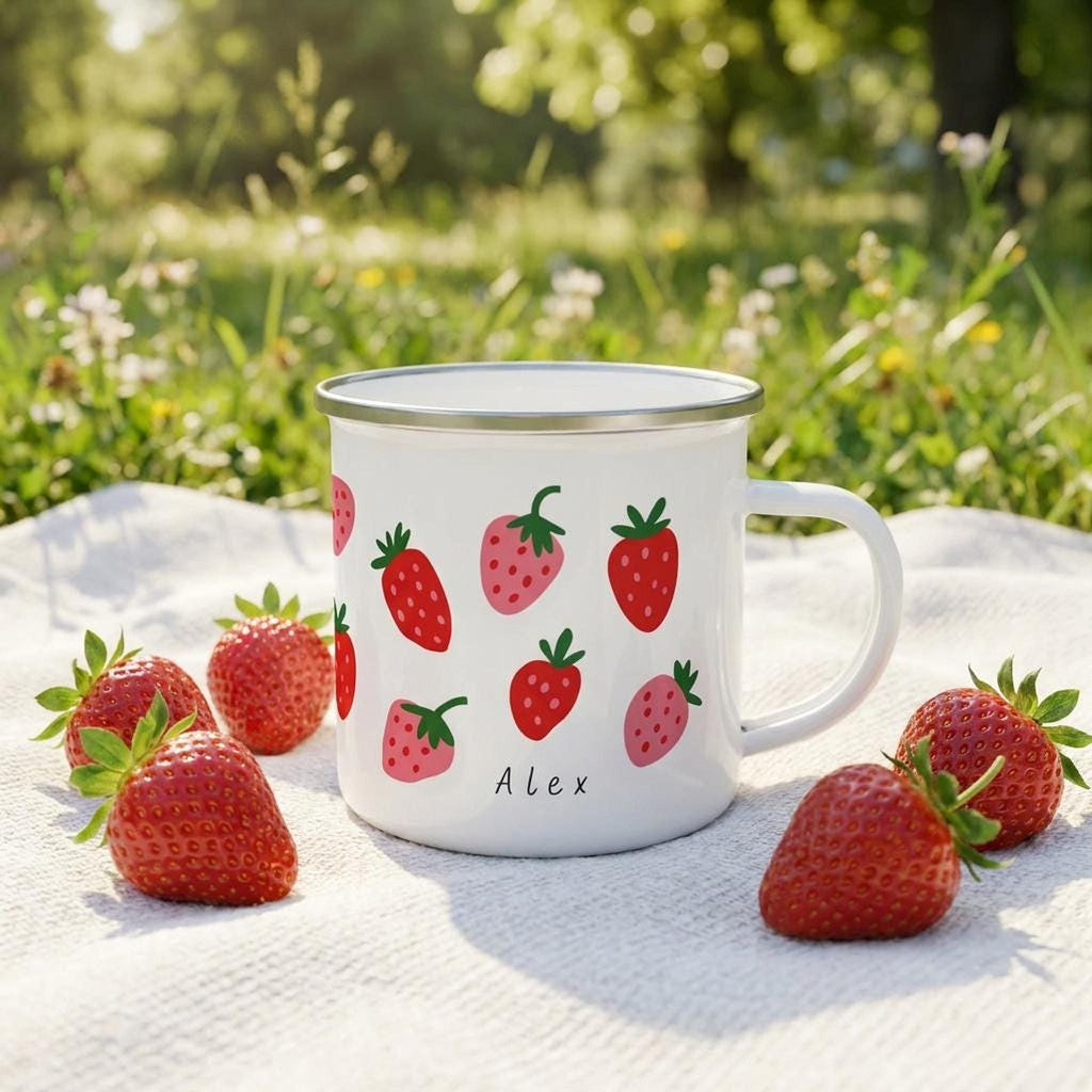 A white enamel mug with a strawberry pattern and the name Alex stands on a white cloth outdoors surrounded by fresh strawberries and green grass.