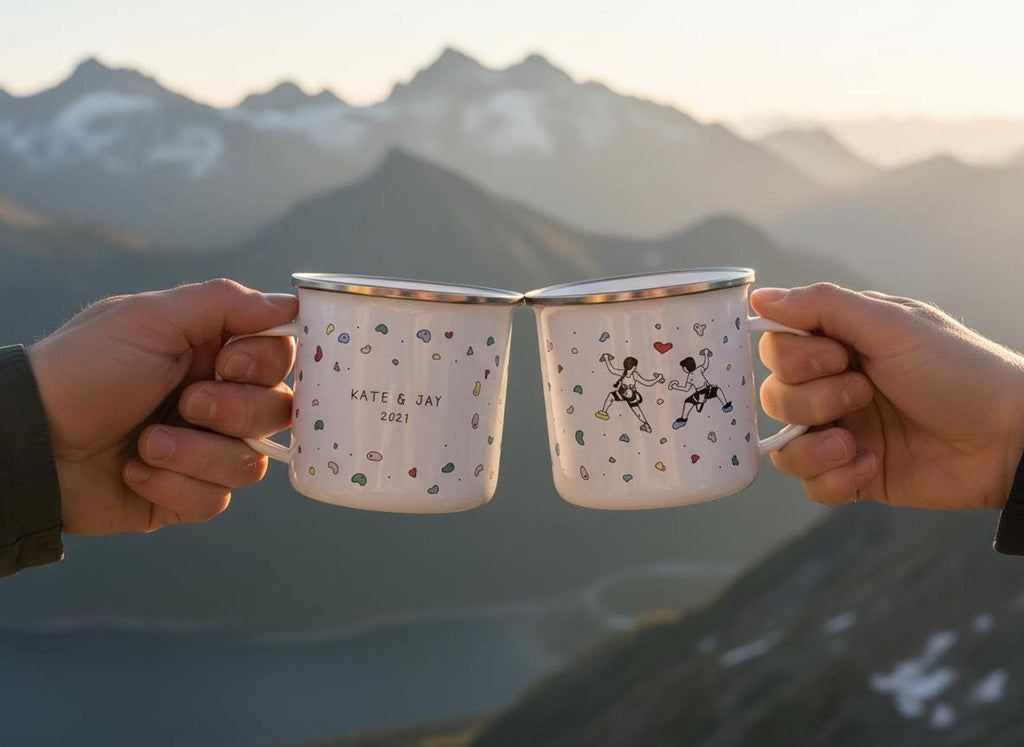 Two hands hold up two white enamel mugs with climbing hold illustrations against a blurry mountain sunset background.