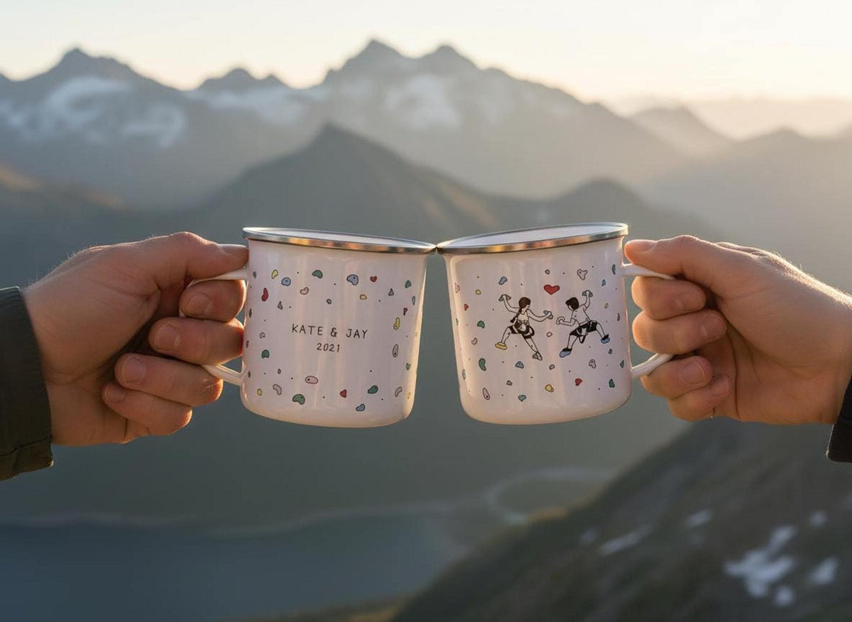 Two hands hold up two white enamel mugs with climbing hold illustrations against a blurry mountain sunset background.