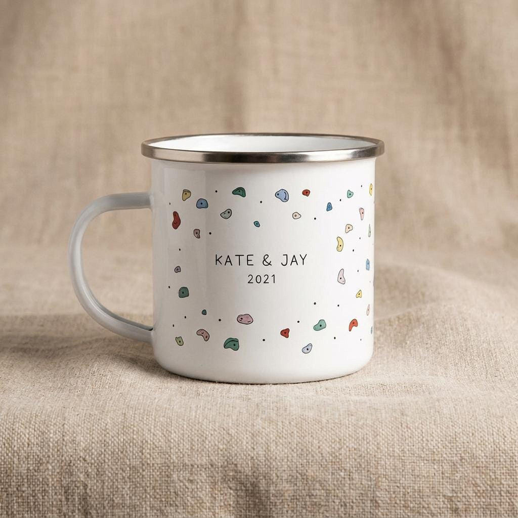 A white enamel mug with colorful climbing hold illustrations and the text KATE AND JAY 2021 on a neutral fabric background.