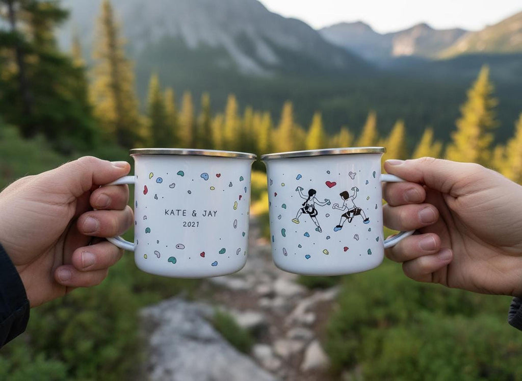 Two hands toast two custom white enamel mugs showing climbing illustrations outdoors with a forested mountain backdrop.