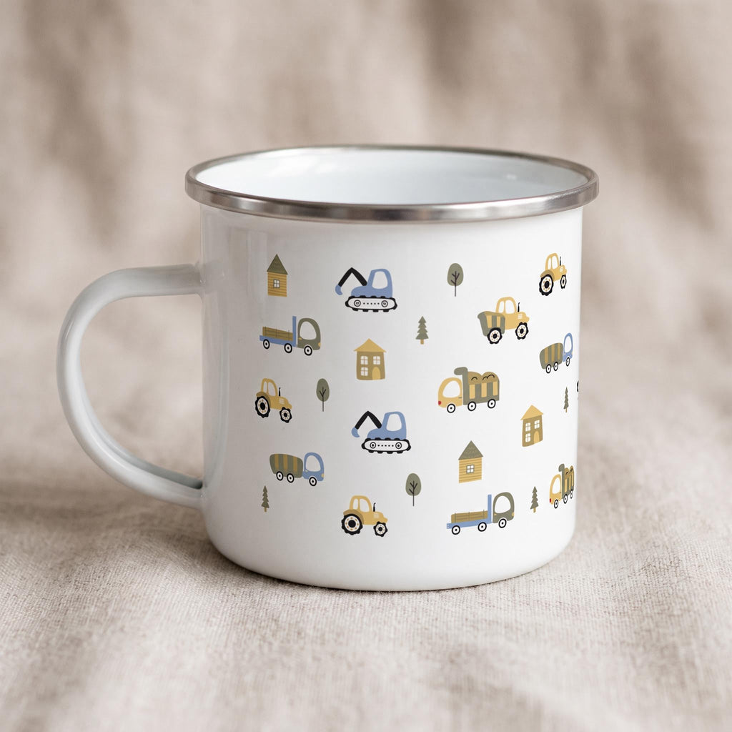 A white enamel mug with a silver rim stands on a neutral fabric surface, decorated with a repeating pattern of cartoon construction trucks, trees, and small houses.