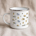 A white enamel mug with a silver rim stands on a neutral fabric surface, decorated with a repeating pattern of cartoon construction trucks, trees, and small houses.
