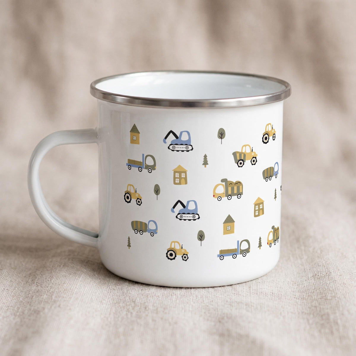 A white enamel mug with a silver rim stands on a neutral fabric surface, decorated with a repeating pattern of cartoon construction trucks, trees, and small houses.