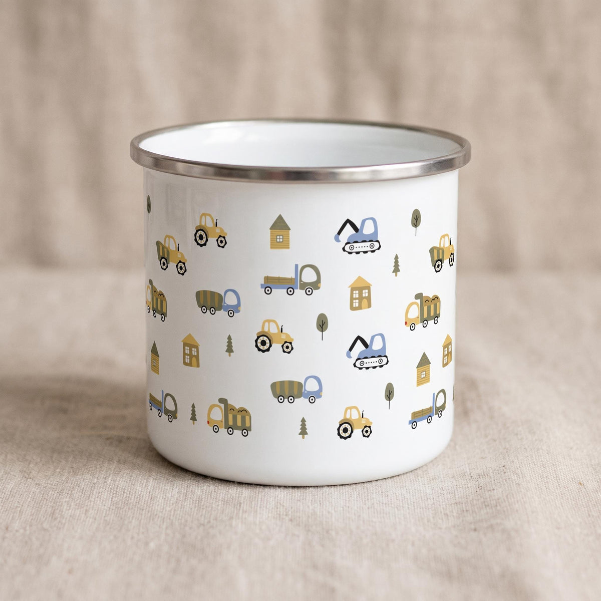 A white enamel mug with a silver rim is angled on a light textured background, displaying a pattern of illustrated construction vehicles, houses, and trees.