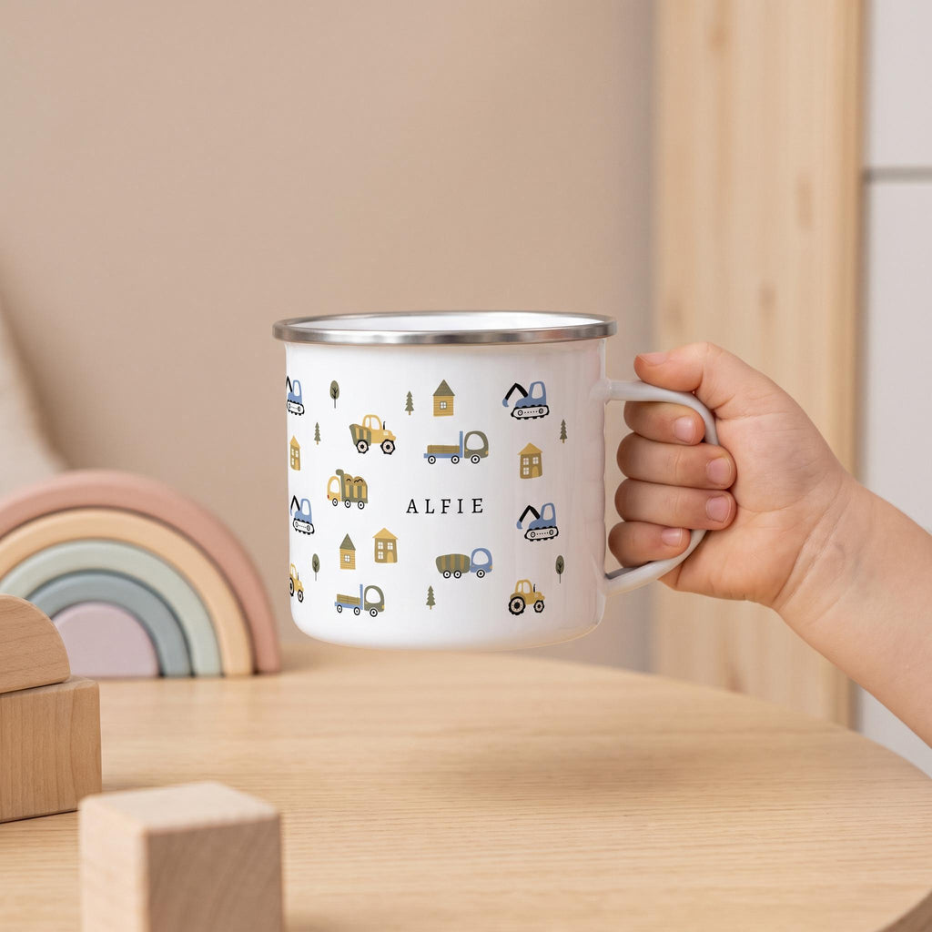 A child holds a personalised white enamel mug featuring a construction vehicle pattern and the name ALFIE, set on a wooden table with wooden toys and a pastel rainbow.