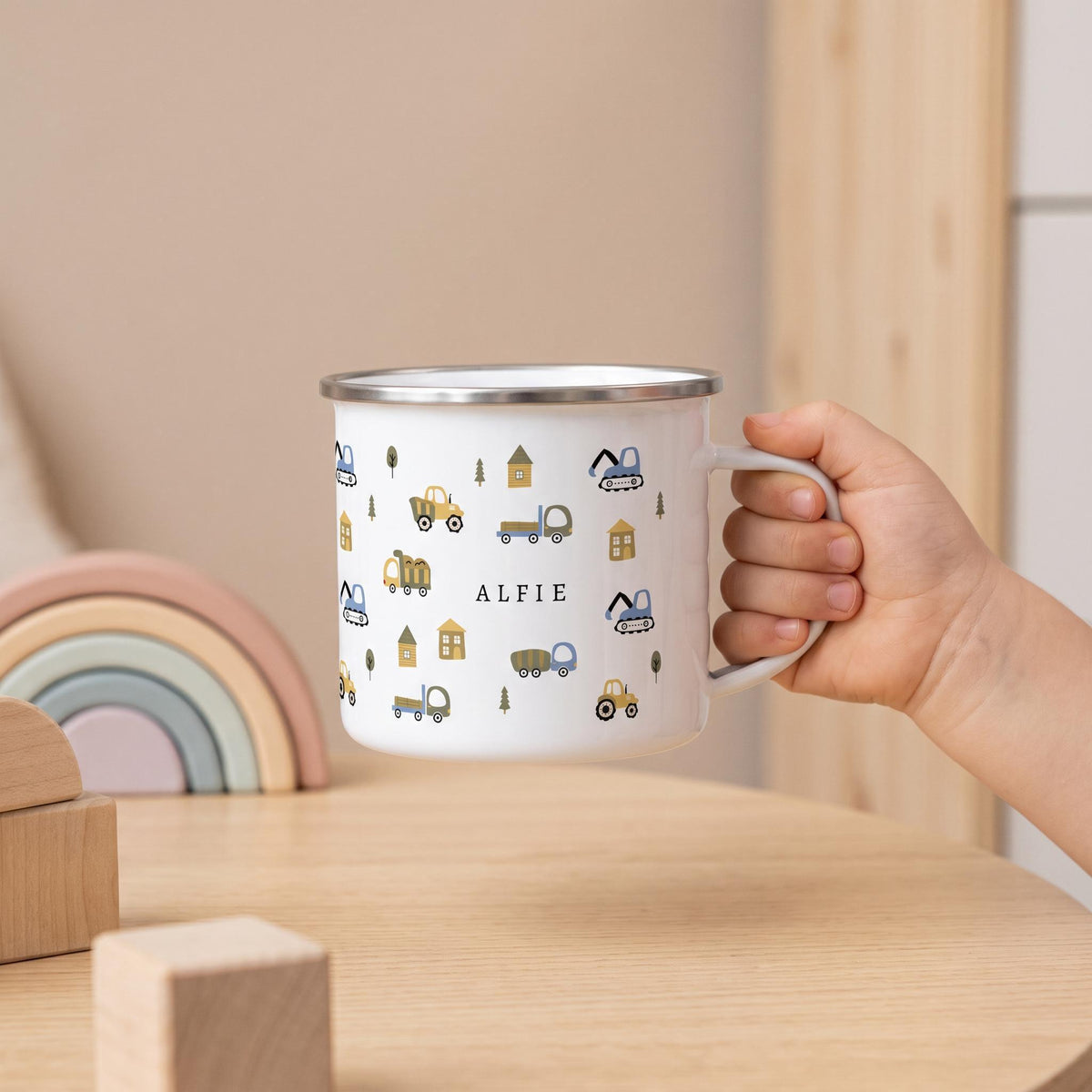 A child holds a personalised white enamel mug featuring a construction vehicle pattern and the name ALFIE, set on a wooden table with wooden toys and a pastel rainbow.