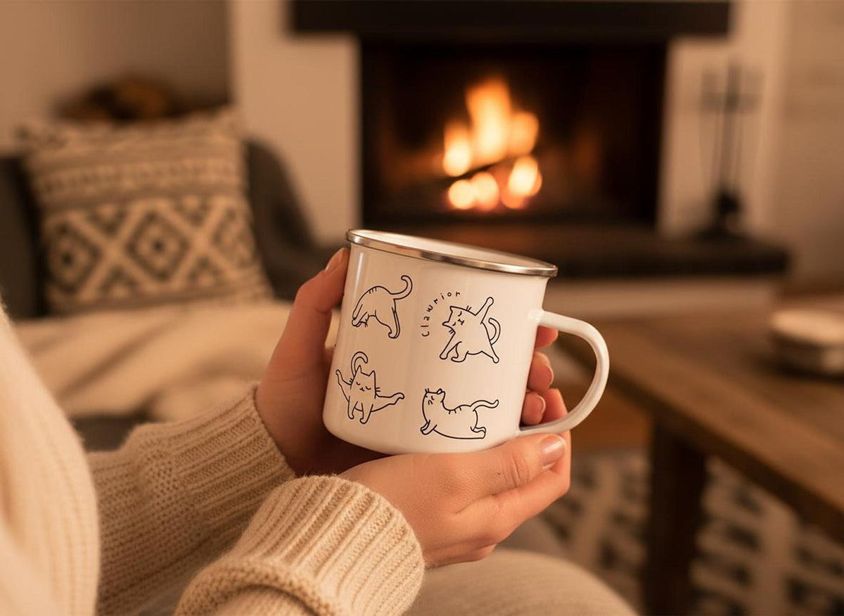 A person holding the cat yoga enamel mug in a cozy room with a lit fireplace in the background.