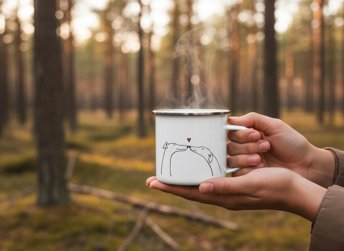 Hands hold a steaming white enamel mug featuring a line drawing of two sighthounds touching noses below a red heart, set against a blurred forest background.