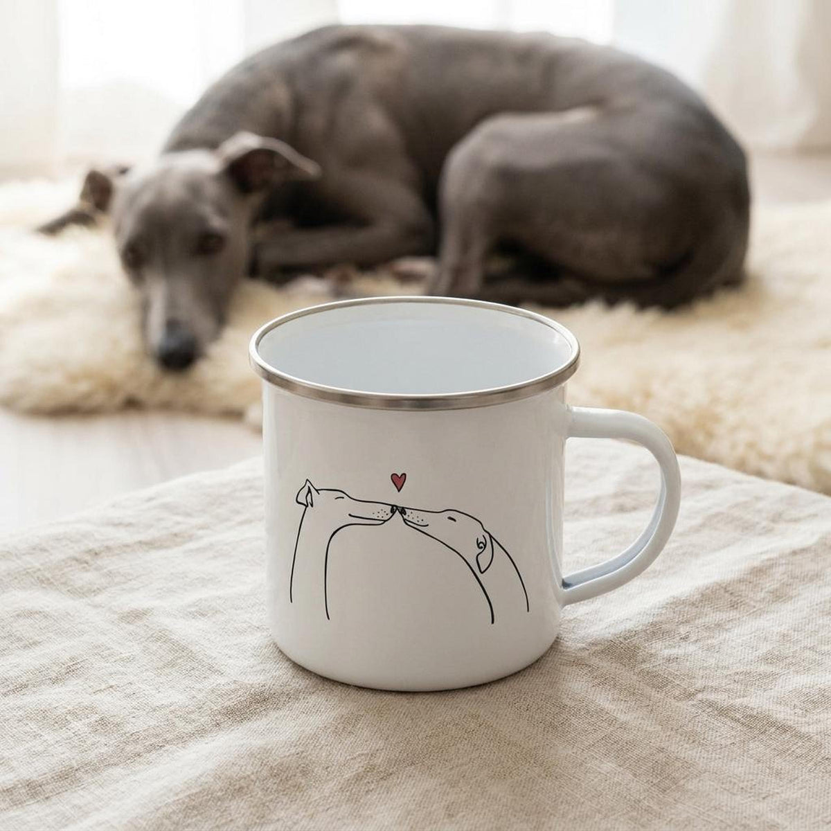 A white enamel mug with a line drawing of two dogs touching noses under a red heart sits on a cloth surface with a grey dog resting in the background.