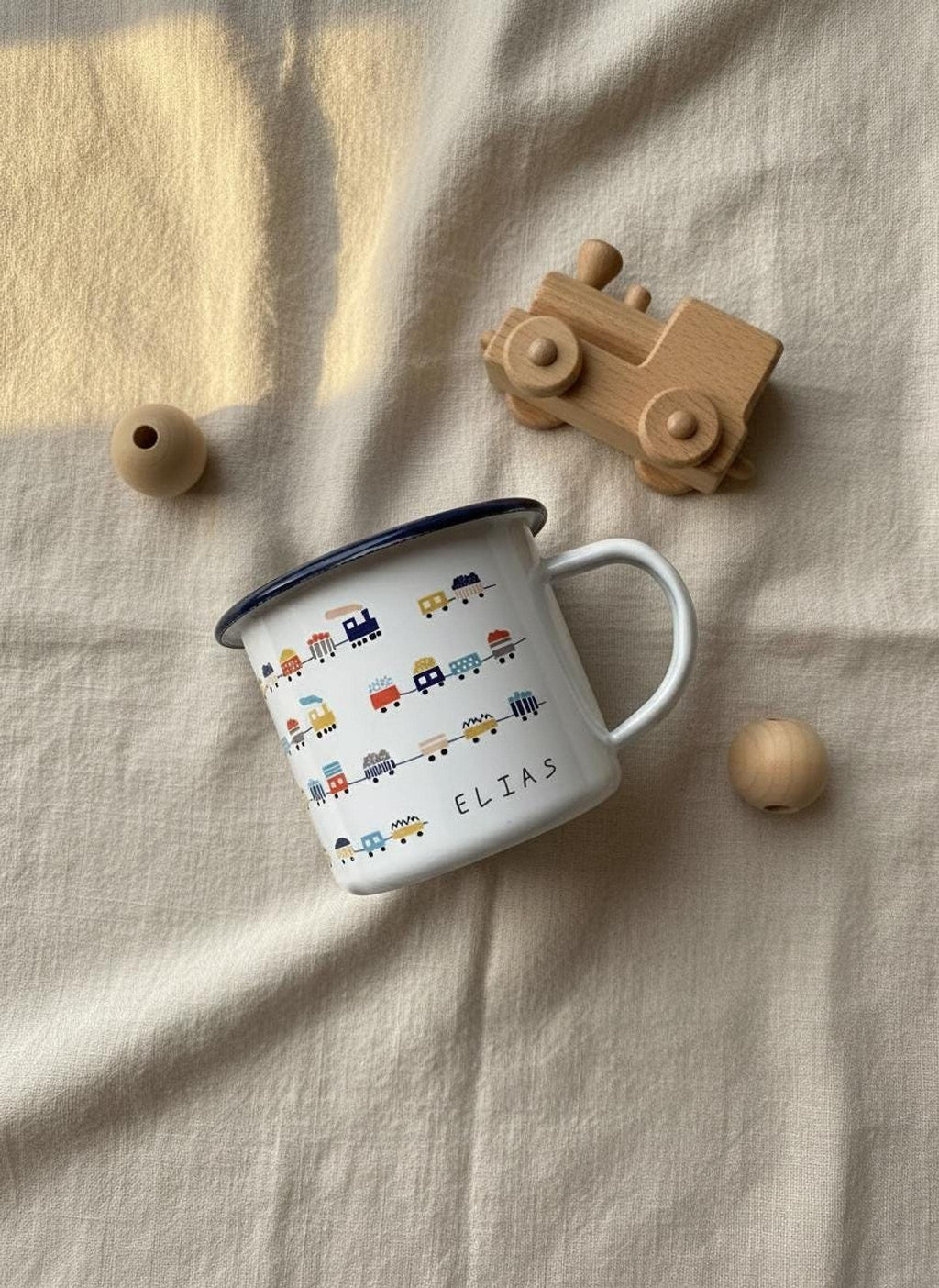 A personalised white enamel mug featuring a pattern of colorful trains and the name Elias, laid on a beige fabric background next to a wooden toy train and beads.