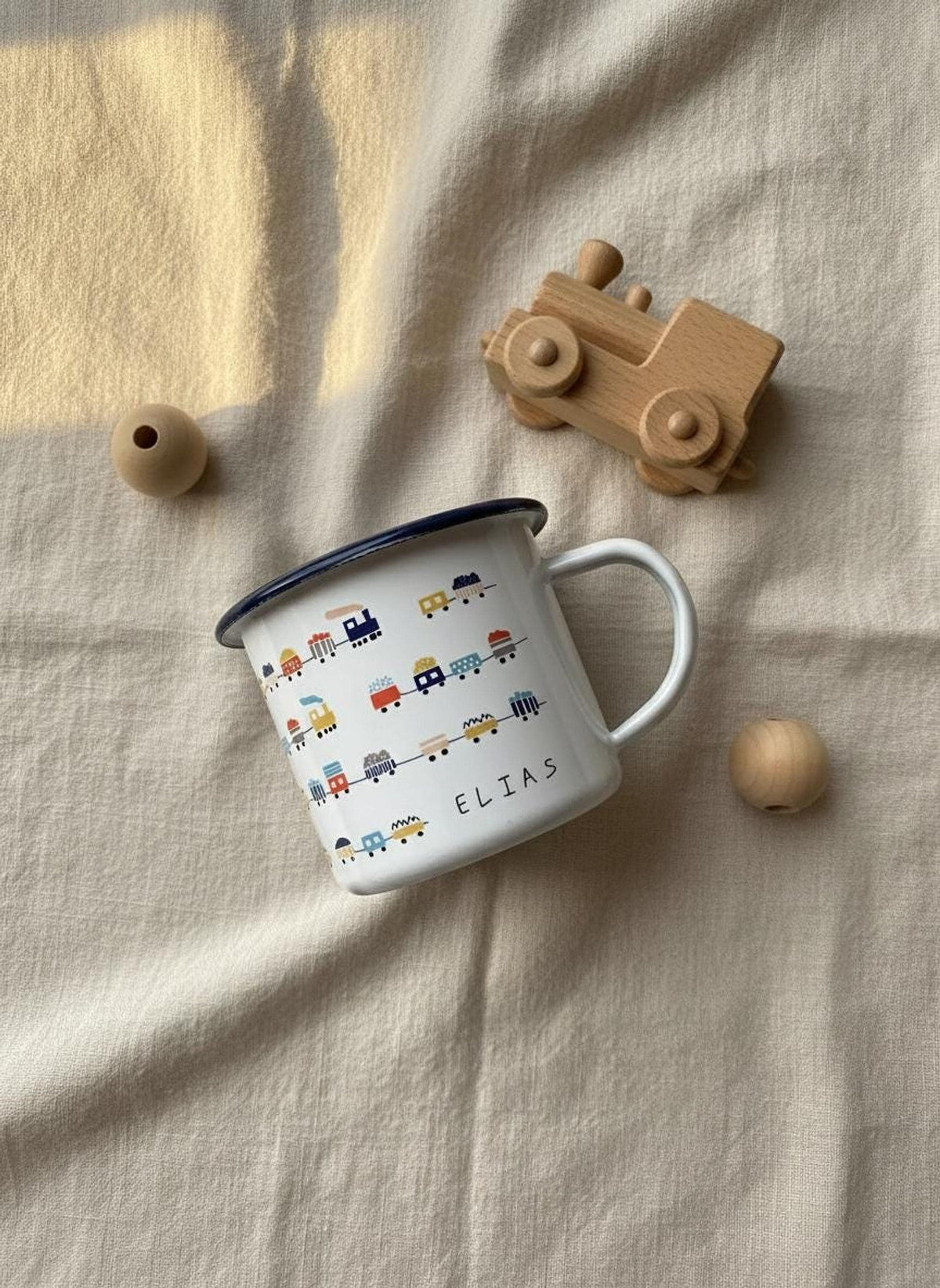 A personalised white enamel mug featuring a pattern of colorful trains and the name Elias, laid on a beige fabric background next to a wooden toy train and beads.