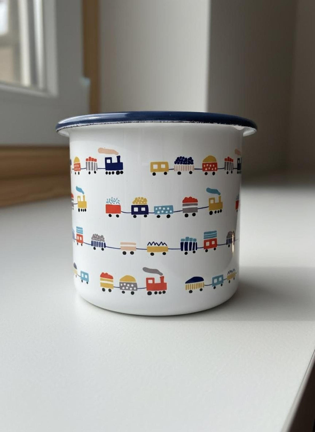 A white enamel mug with a blue rim and multiple rows of printed colorful trains sitting on a white surface.