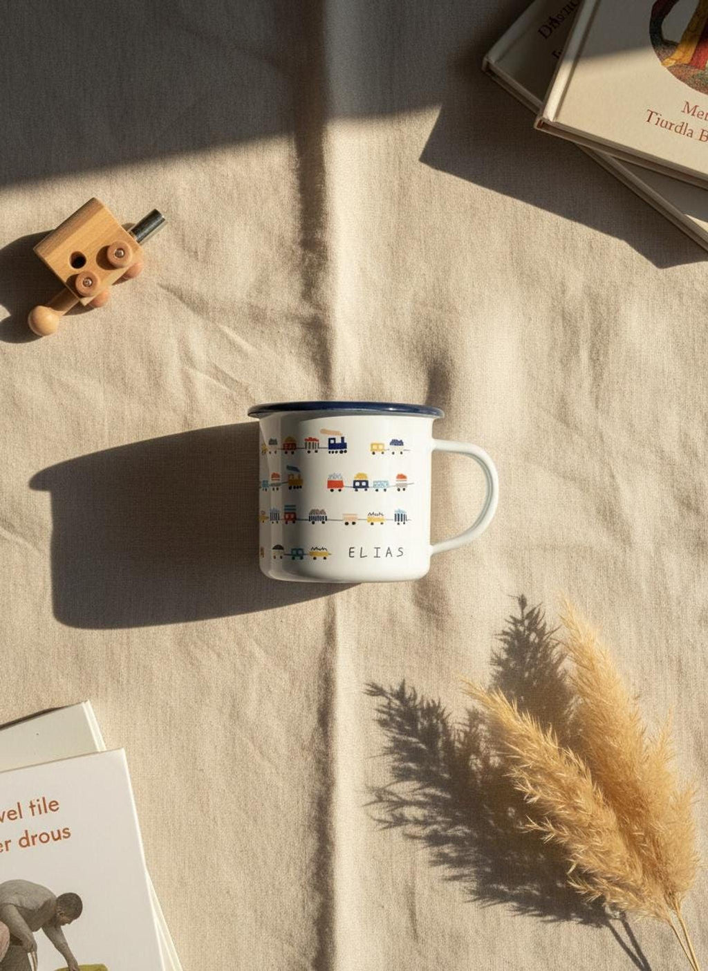 A white enamel mug with a train pattern and the name Elias is displayed on a textured fabric surface surrounded by books, a toy, and dried grasses.