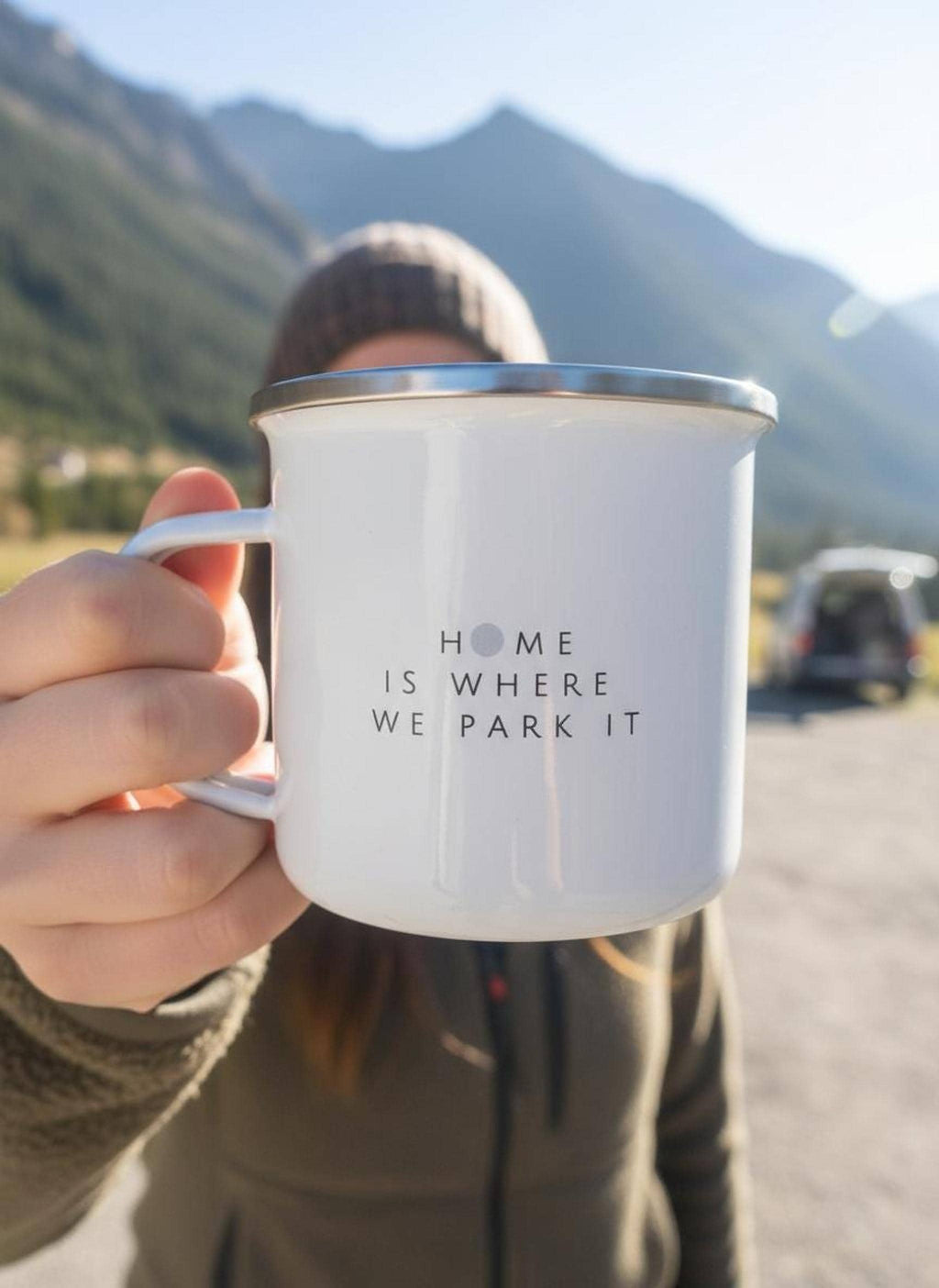 A person holds up a white enamel mug printed with the text Home Is Where We Park It with a mountain landscape in the background.