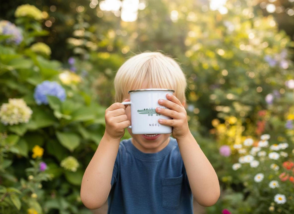 A child holding a white enamel mug with a crocodile illustration and the name Noel in a green garden setting.