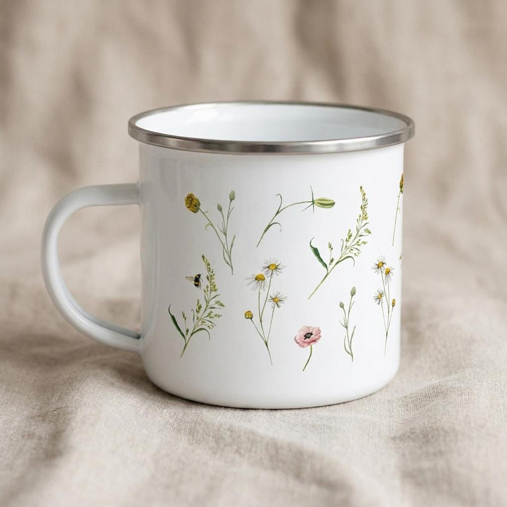 A white enamel mug with a silver rim sits on a beige fabric surface, printed with botanical illustrations of wildflowers and bees.