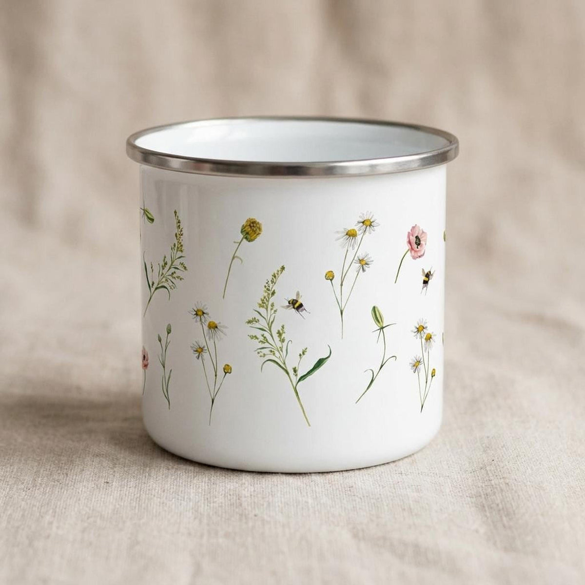 A side view of a white enamel mug with a silver rim, decorated with watercolor-style wildflowers and flying bees on a textured neutral background.