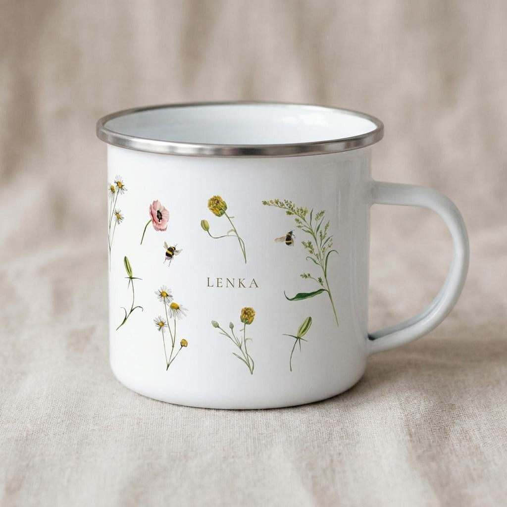 A white enamel mug featuring illustrations of assorted wildflowers, bees, and the custom text LENKA rests on a neutral fabric backdrop.
