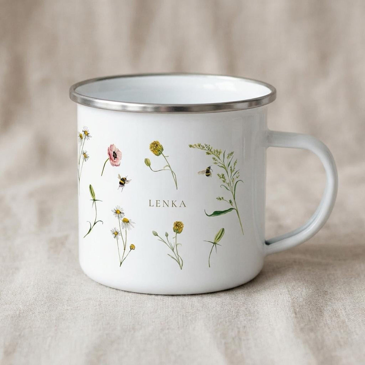 A white enamel mug featuring illustrations of assorted wildflowers, bees, and the custom text LENKA rests on a neutral fabric backdrop.