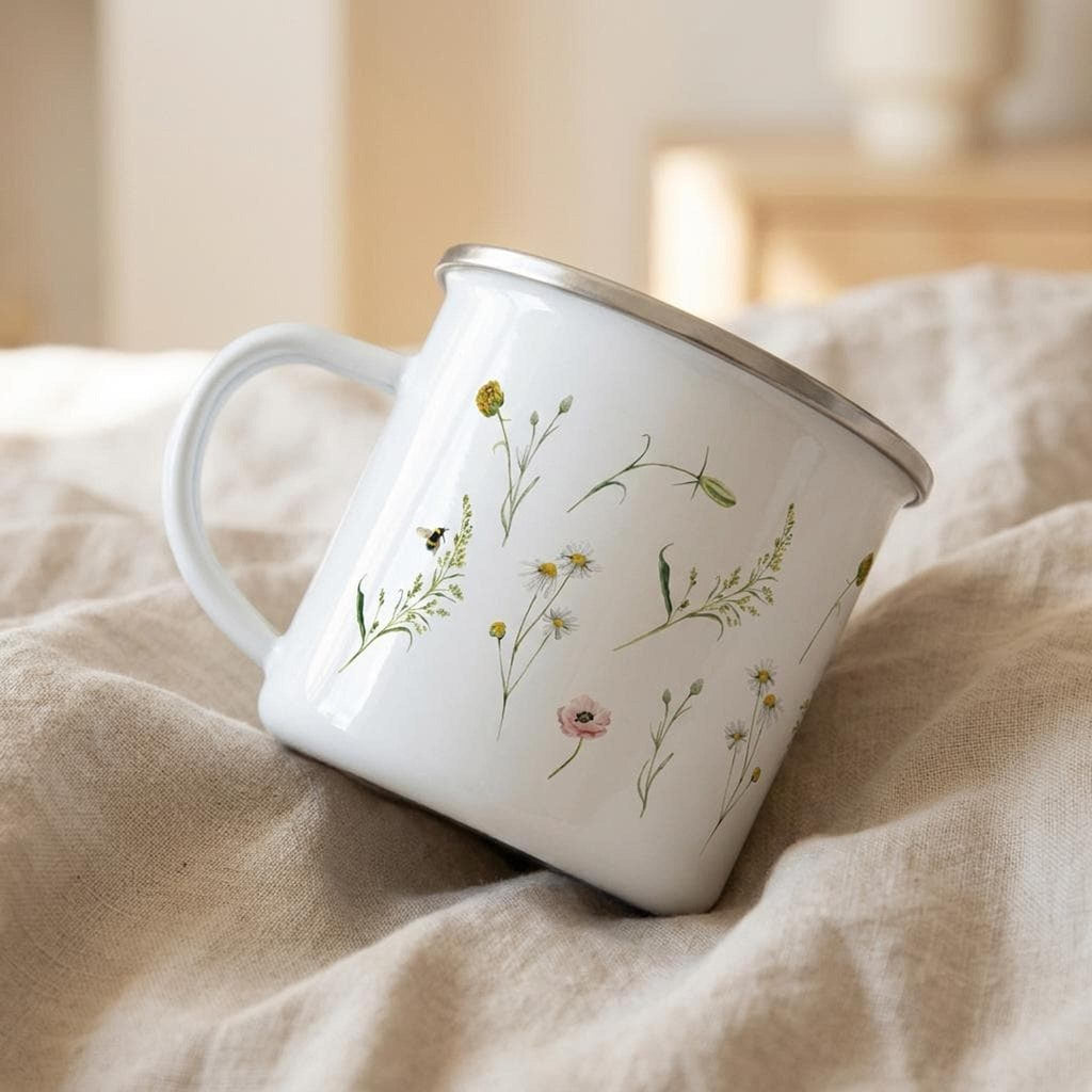 A white enamel mug with wildflowers printed on its surface rests at an angle on folded beige fabric.