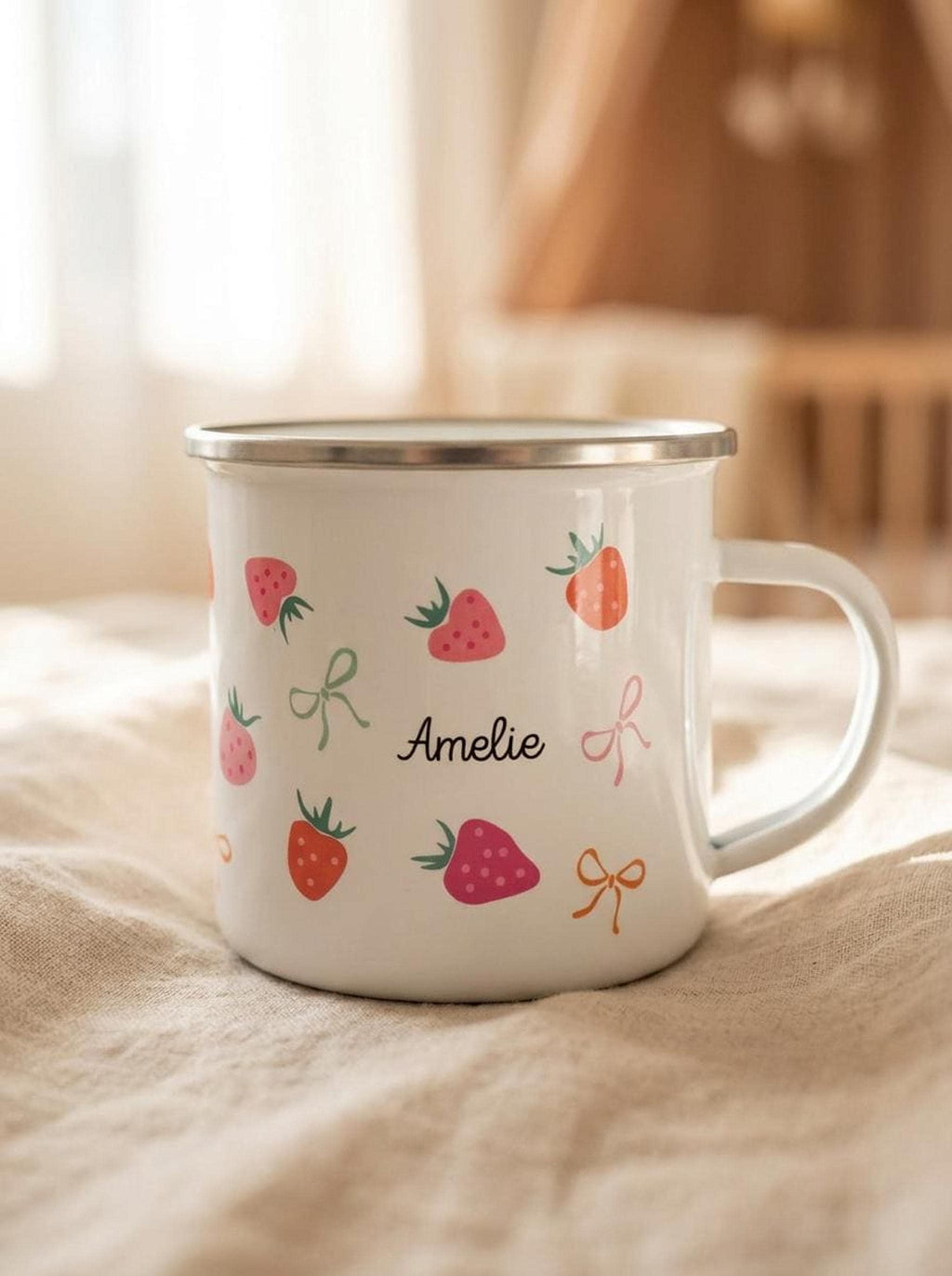 A white enamel mug with a silver rim, printed with strawberries, bows, and the name Amelie, resting on a beige cloth against a soft, sunlit room background.